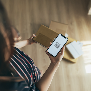 A person is holding a smartphone with a QR code in their hand while standing next to several boxes.