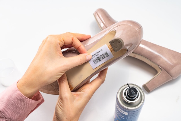 A hand is holding a shoe with a price tag, while a spray can stands next to it. The shoe is a beige high heel.