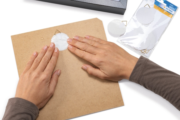 Hands are pressing a round hanger from HERMA onto a piece of brown cardboard, while a pack of additional stickers sits beside it.