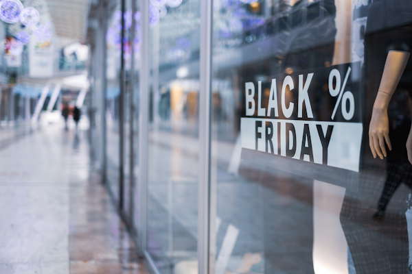 Store window with a 'BLACK FRIDAY' sign displaying discounts, in a shopping mall. People are walking by in the background.