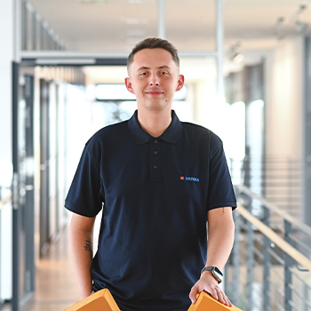 A young man in a dark blue polo shirt stands in a modern office hallway, holding an orange object in his hand.