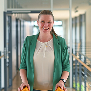 A woman with long hair is wearing a green blazer and standing in a modern hallway. She is smiling and has a watch on her wrist.