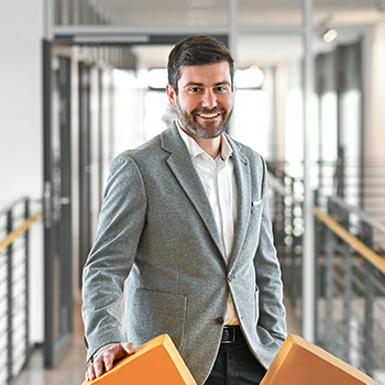 A man in a gray blazer smiles while holding two orange objects in a modern office setting.