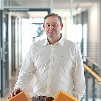 A man in a white shirt is holding two orange boxes in a modern office with large windows.
