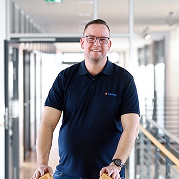 A man with glasses and a short haircut is wearing a dark blue polo shirt and standing in a modern office hallway.