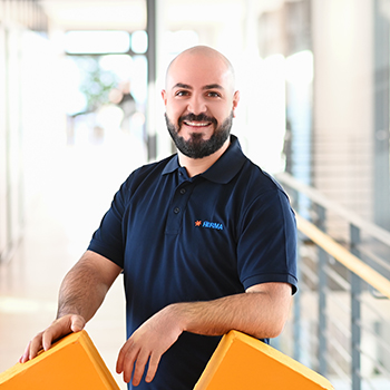 A man with a beard is wearing a black polo shirt and standing in a modern interior while holding yellow objects in both hands.
