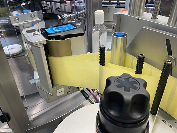 A labelling device with a yellow label, which is applied to bottles, is located in a production environment.
