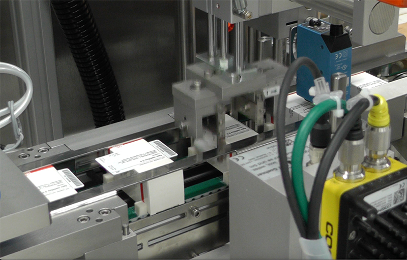 Automated machine components that apply labels to products. Visible labels with text on a conveyor belt.