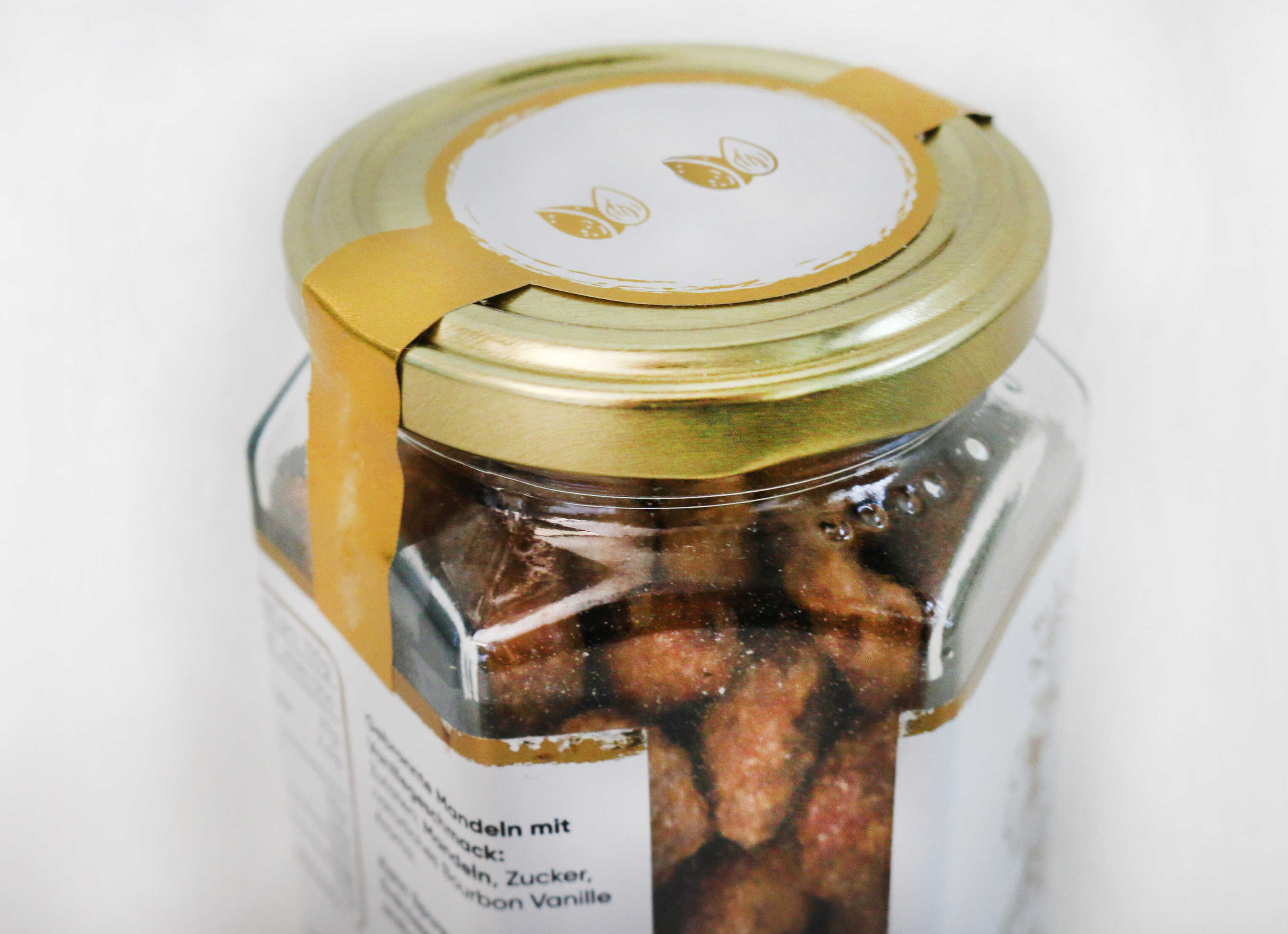 A glass with a gold-colored lid, filled with roasted almonds. On the lid, there is a label with a minimalist design.