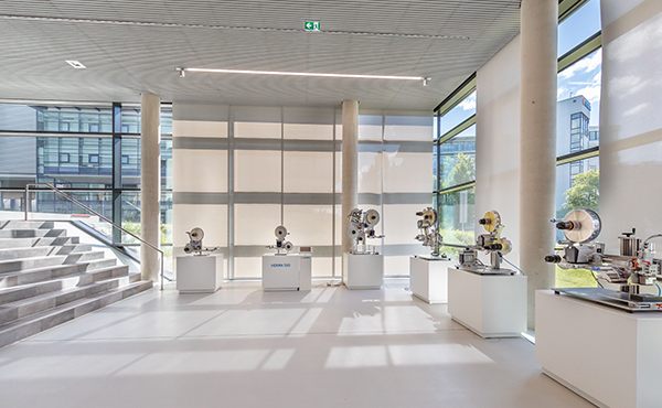 Modern machines and equipment are displayed on white pedestals in a bright, airy room with large windows.