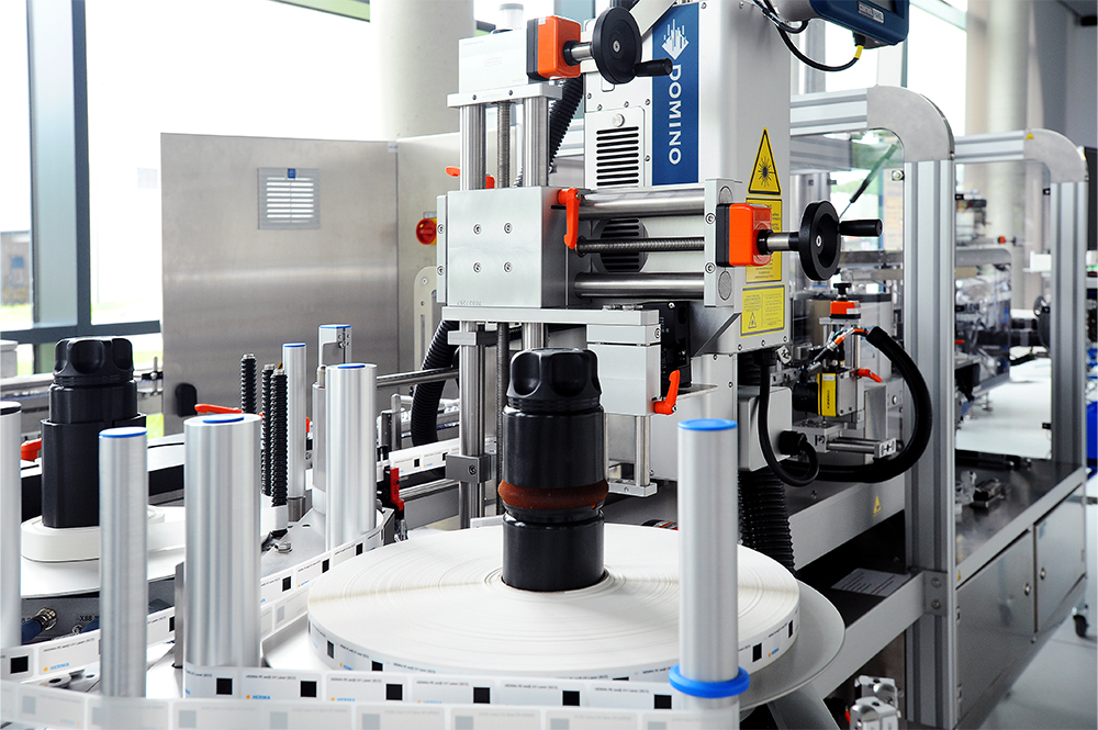 An automated packaging machine with bottles on a conveyor belt, equipped with labelling and printing systems.