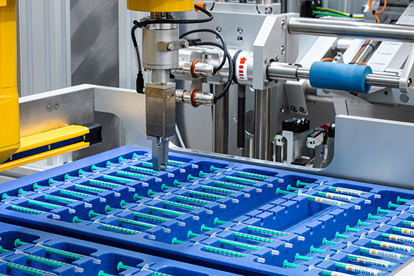 Automated machine that processes syringes in blue containers, with a robotic arm that picks up a syringe.