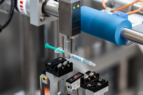 An automated gripper holds a syringe labelled 'HERMA' in an industrial setting.