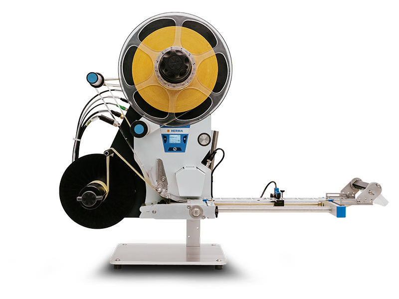 A labelling machine with a large spool that applies yellow labels on a conveyor belt. It has several connections and controls.