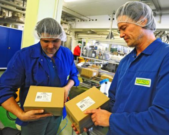 Two workers in blue overalls and hairnets are holding boxes in a production environment and checking their contents.