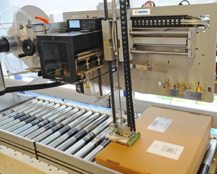 Automated labelling system with a box on a conveyor belt, equipped with printing and labelling mechanisms.