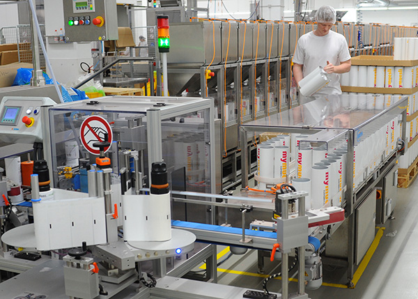 A worker in a manufacturing facility is filling containers at an automated bottling line. Machines and packaging are visible in the background.