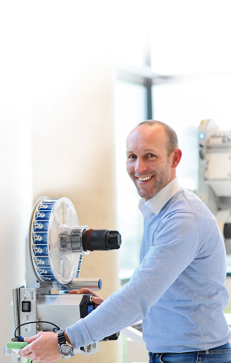 A man in a bright sweater is operating a technical device with a large coil. He is smiling warmly at the camera.