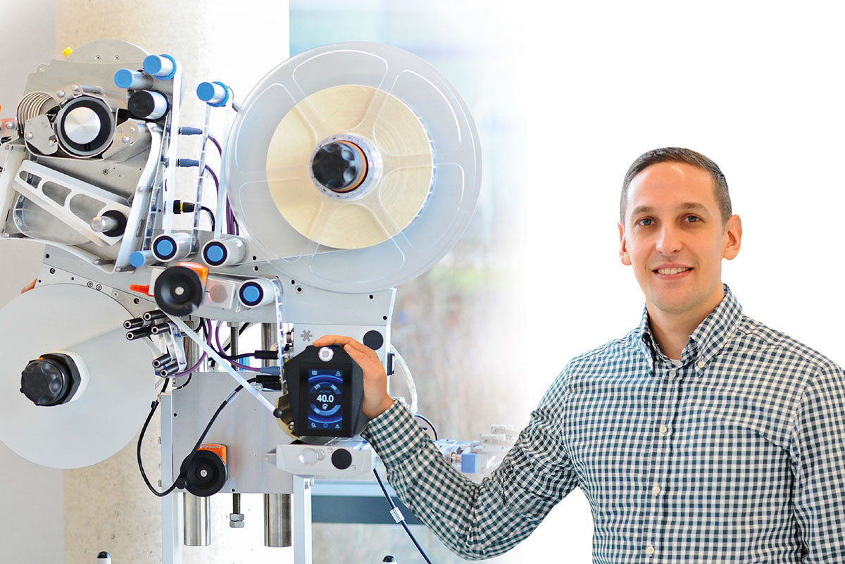 A man stands next to a modern labelling machine, equipped with various rolls and control elements.