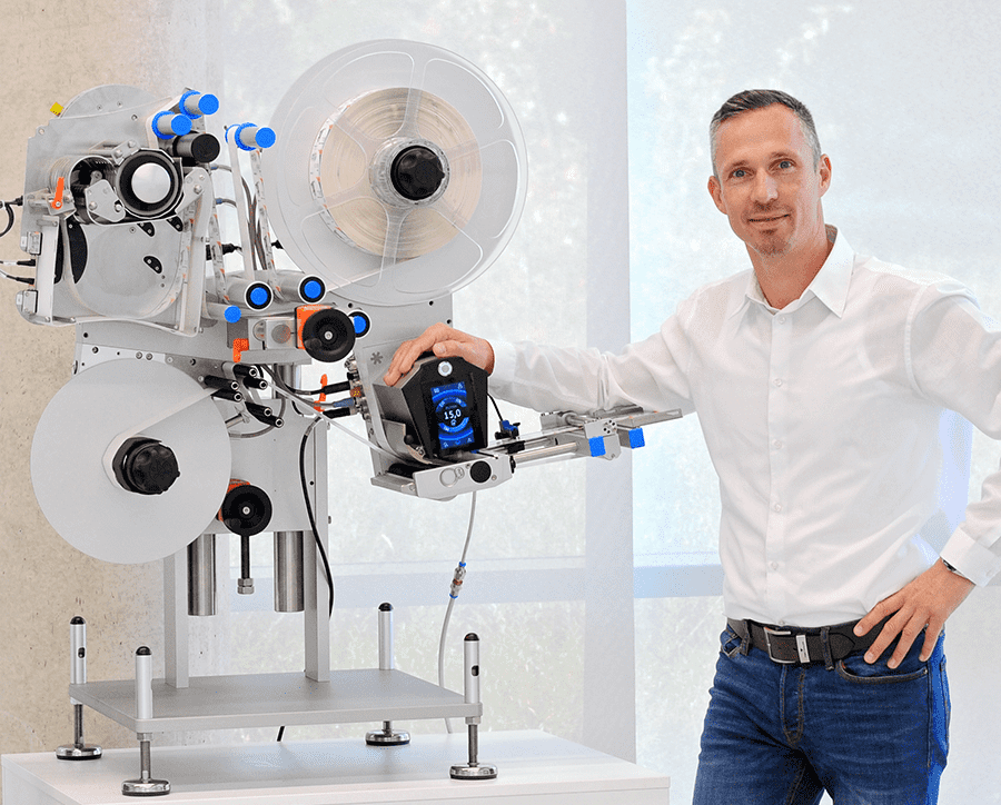 A man stands next to a modern labelling machine, holding a control device in his hand. The machine has several rollers and control elements.