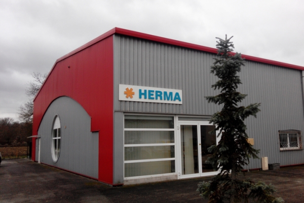 Facade of a modern building with a red roof and gray walls, labelled with 'HERMA.' A tree stands in the foreground.