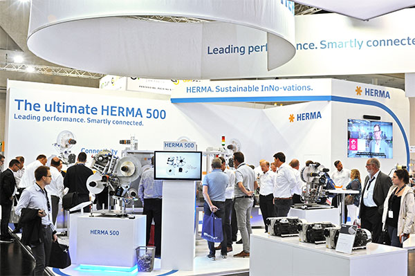 HERMA's trade show booth featuring the HERMA 500. Visitors interact with the displayed machines and information.