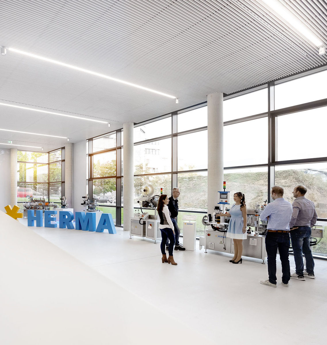 A group of five people stands in a modern room with large windows, surrounded by machines. A large blue HERMA sign is visible.