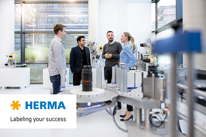 Four people are discussing in a modern room, surrounded by machines and equipment for labelling. A HERMA logo is visible.