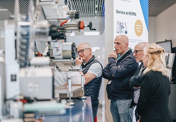 Four people are attentively watching a machine demonstration at a trade fair, while information about the service at HERMA is being displayed.