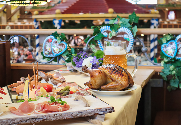 A table with a selection of sausages, cheese, and vegetables, accompanied by a roasted chicken and a large beer glass in a festive atmosphere.