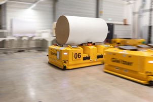 Yellow autonomous transport carts are moving around in a warehouse, one of them carrying a large roll of paper.