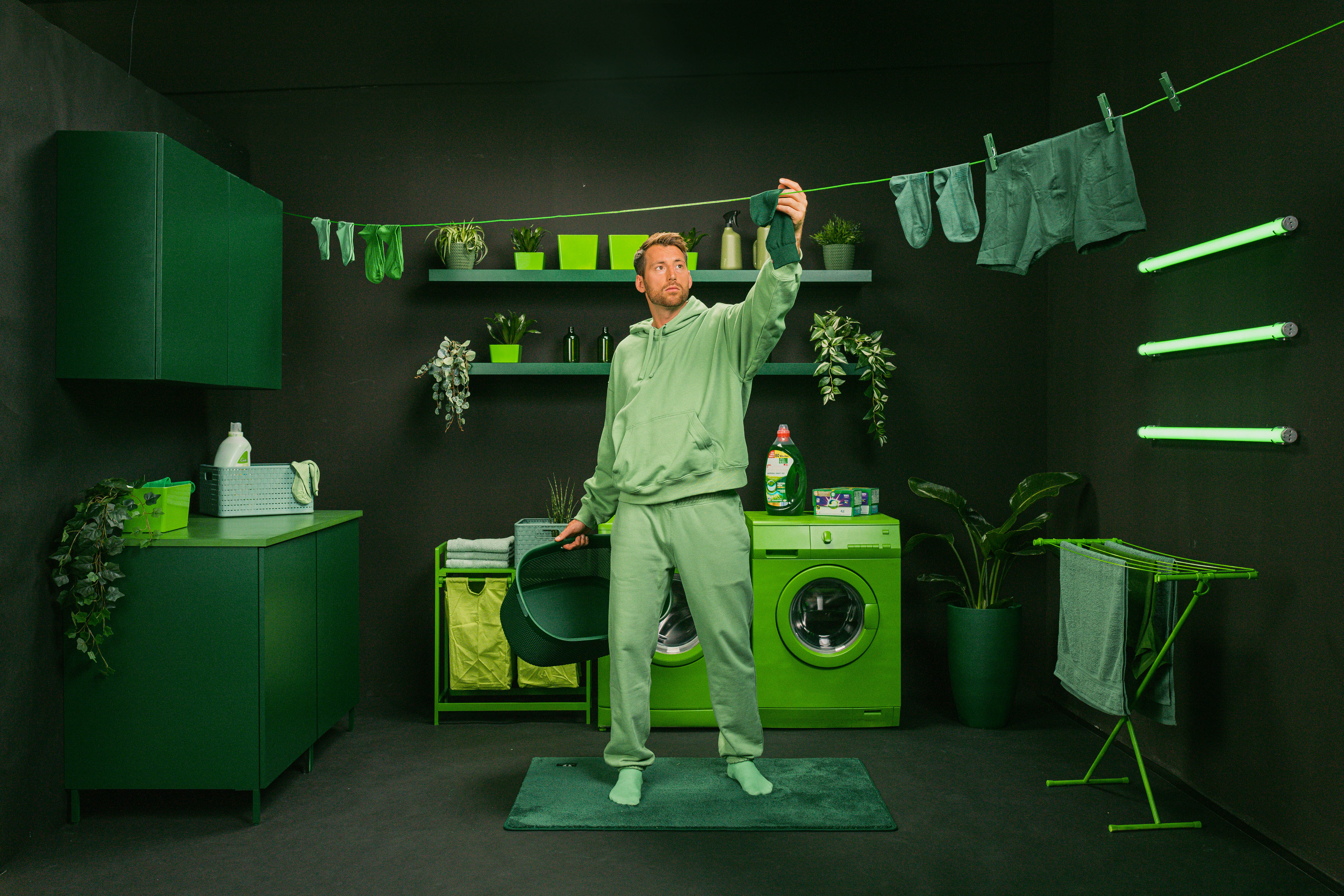A man in green clothing is hanging laundry in a monochromatic green room. A washing machine and plants are visible in the background.