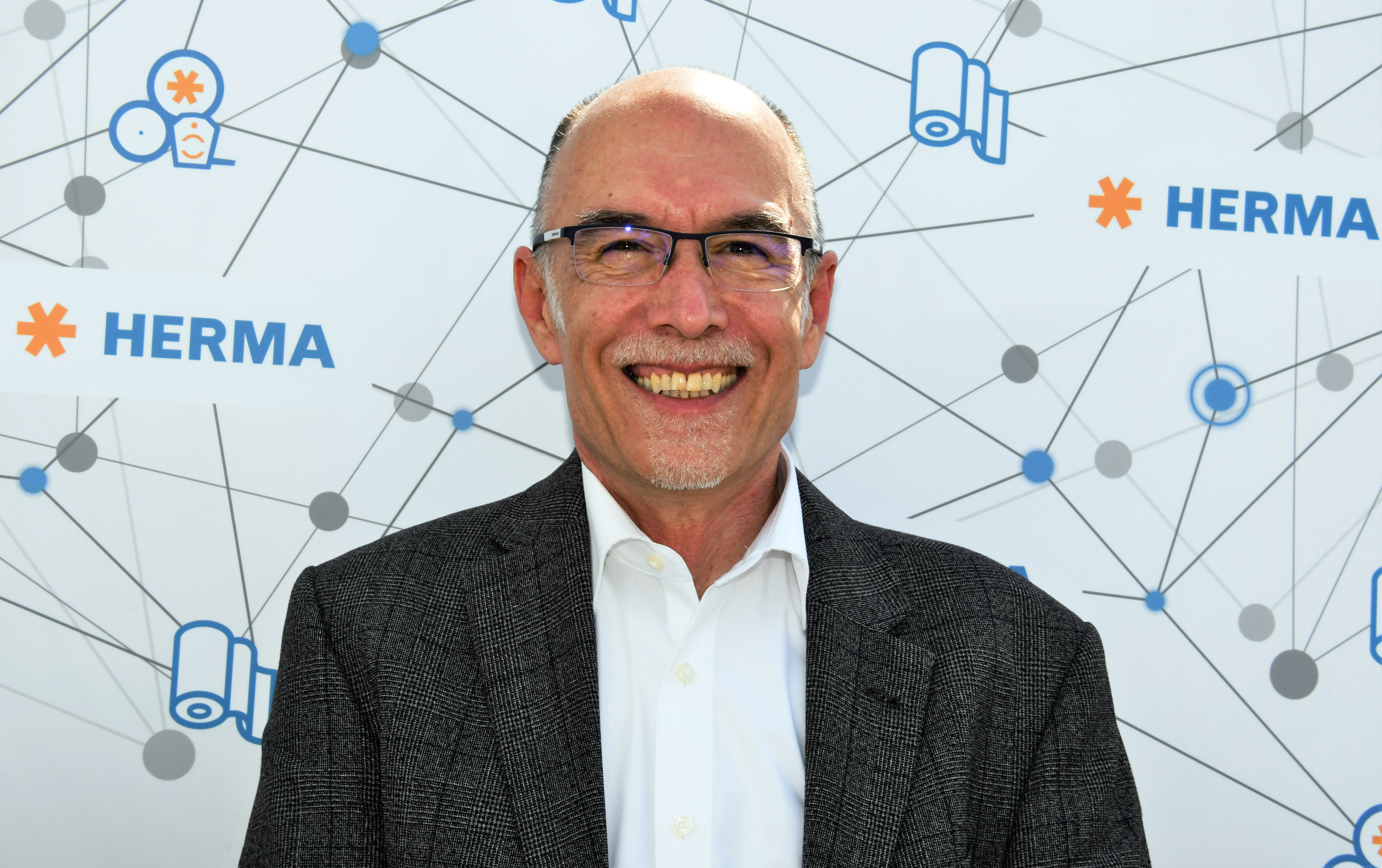 A man in glasses and a gray suit smiles against a background featuring the HERMA logo and graphic elements.