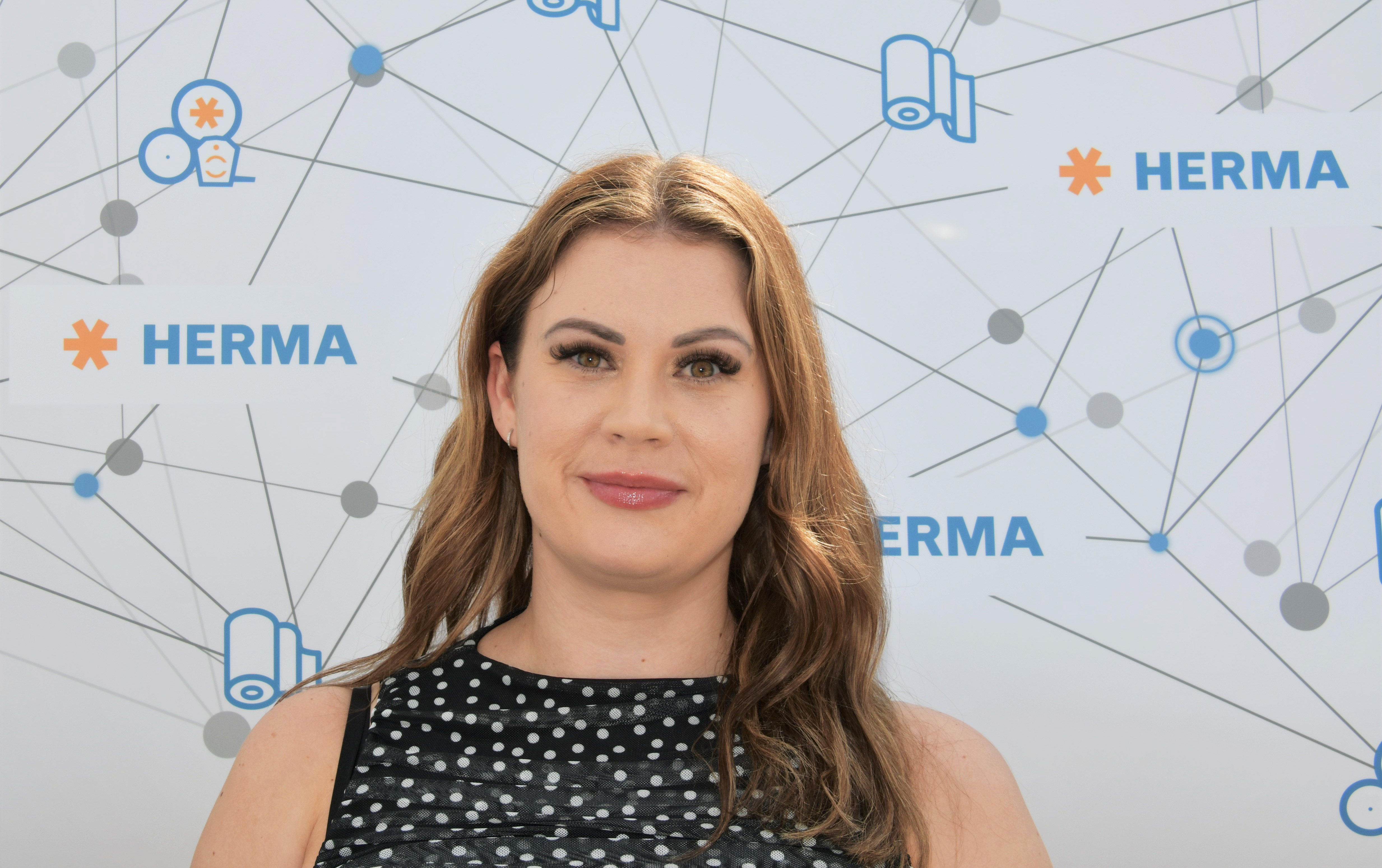 A woman with long brown hair and a polka-dotted dress stands against a background featuring the HERMA logo and graphic elements.