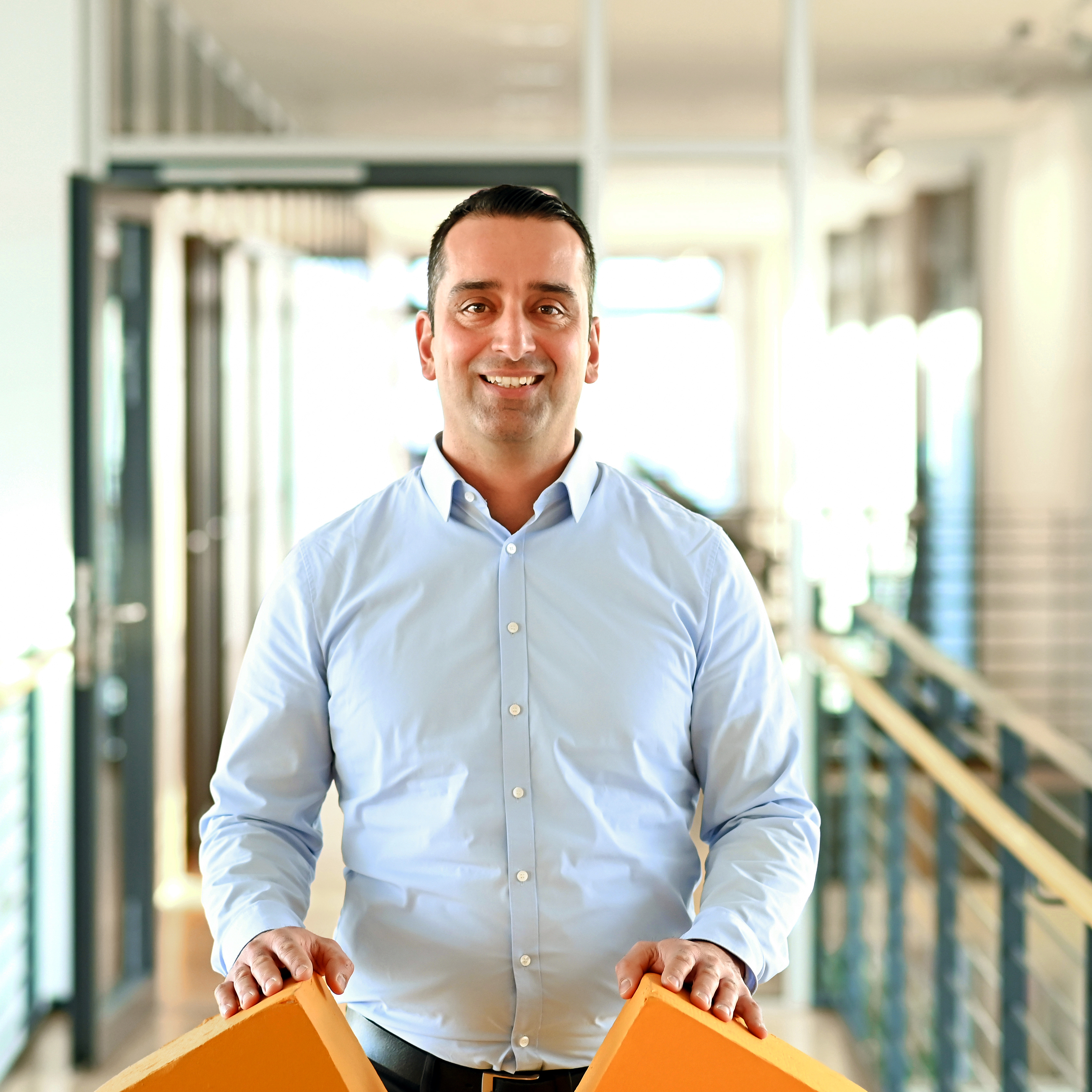 A man in a light blue shirt stands in a modern office, holding two orange dice in his hands.
