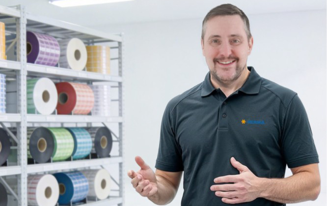 A man in a black polo shirt stands in front of a shelf filled with colorful rolls. He is smiling and gesturing warmly.