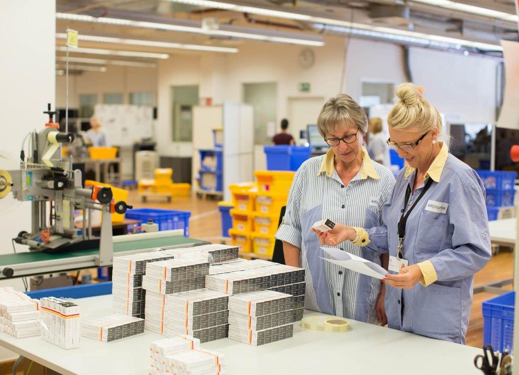 Two women in work attire are checking documents and packages at a table in a production environment.