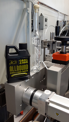 A bottle and a bottle of grill and BBQ sauce are sitting on a machine surface in a production environment.