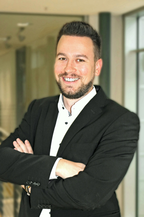 Man in a black suit with crossed arms, smiling warmly in a modern interior.