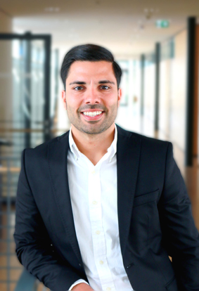 A man in a black suit and white shirt smiles as he stands in a modern office hallway.