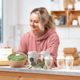 A woman in a pink hoodie is preparing healthy meals, including a bowl of salad and several coffee mugs on a table.