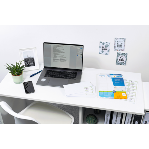 Ein Schreibtisch mit einem Laptop, einem Smartphone, einer Pflanze und verschiedenen Dokumenten. An der Wand hängen dekorative Notizen.