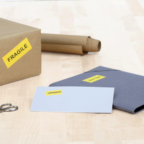 Ein brauner Karton mit dem Aufkleber "FRAGILE", daneben ein graues Kuvert mit dem Aufkleber "URGENT!" und eine Schere auf einem Tisch.