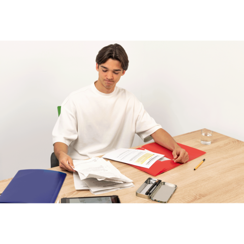 Ein junger Mann sitzt an einem Tisch, hält Dokumente in der Hand und hat verschiedene Ordner sowie ein Tablet vor sich.