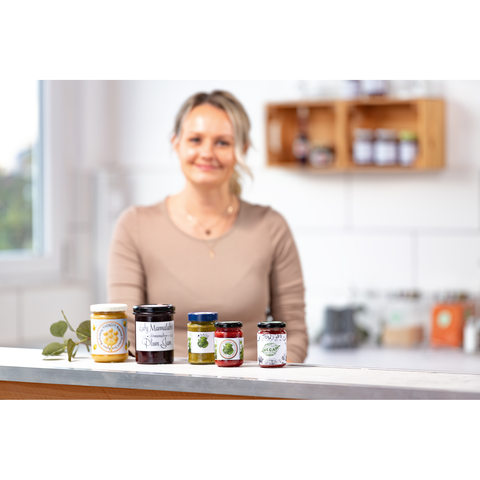 Eine Frau steht an einer Küchenarbeitsplatte mit verschiedenen Marmeladengläsern. Im Hintergrund sind Regale mit weiteren Lebensmitteln sichtbar.