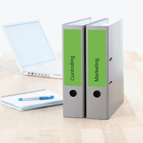 Zwei Ordner mit den Beschriftungen "Controlling" und "Marketing" stehen auf einem Tisch neben einem Laptop und einem Notizblock.