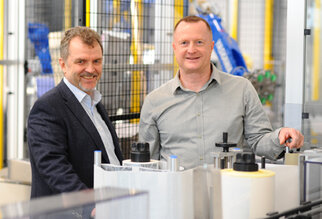 Two men are standing in a modern production environment, surrounded by machines and equipment, smiling at the camera.