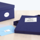 Eine geöffnete, dunkelblaue Geschenkbox mit der Aufschrift „Happy Birthday!“ und zwei Umschlägen in hellblau und grau daneben.
