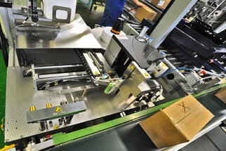 Automated packaging machine with conveyor belts and boxes in the background. Various mechanical components are visible.