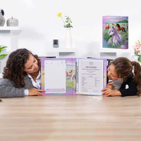 Two girls are sitting at the table, looking at an open report card in a colorful binder featuring a unicorn design.